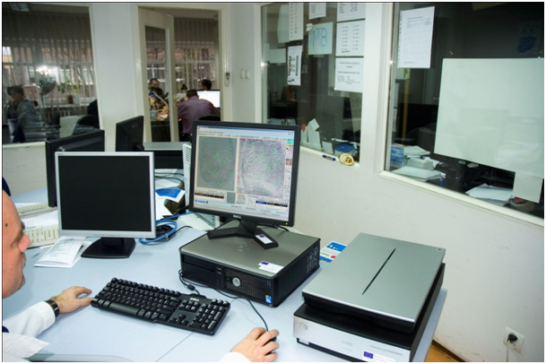 Ilustracja przedstawiajaca pracownię badań daktyloskopijnych. Na pierwszym planie widnieją komputery stojące na biurku